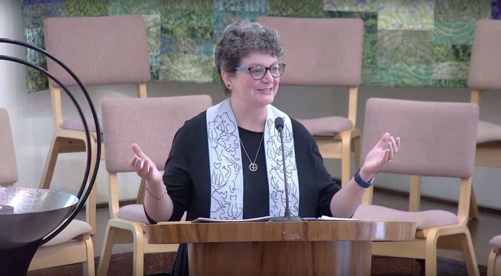 Rev. Mary Gear at the pulpit, wearing a patterned, white stole and chalice necklace, gesturing to the congregation during a Sunday Service