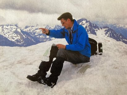 Jim Knudson, sitting on a snowy mountaintop, looking at a map and pointing off into the distance