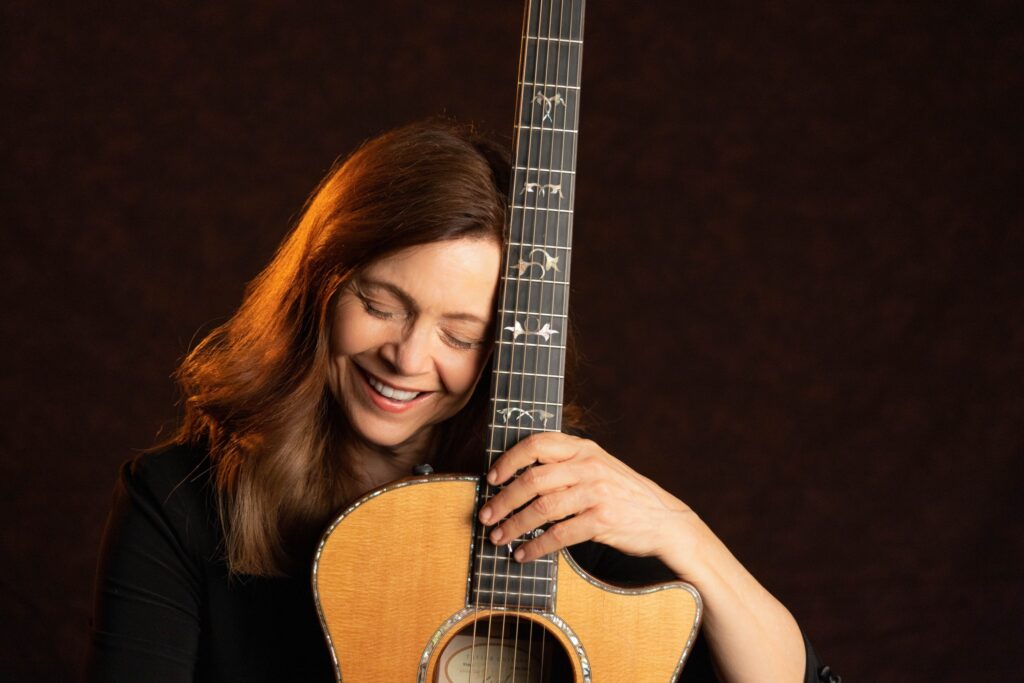 a middle-aged white woman with long brown hair, smiling, eyes closed, posing with a guitar
