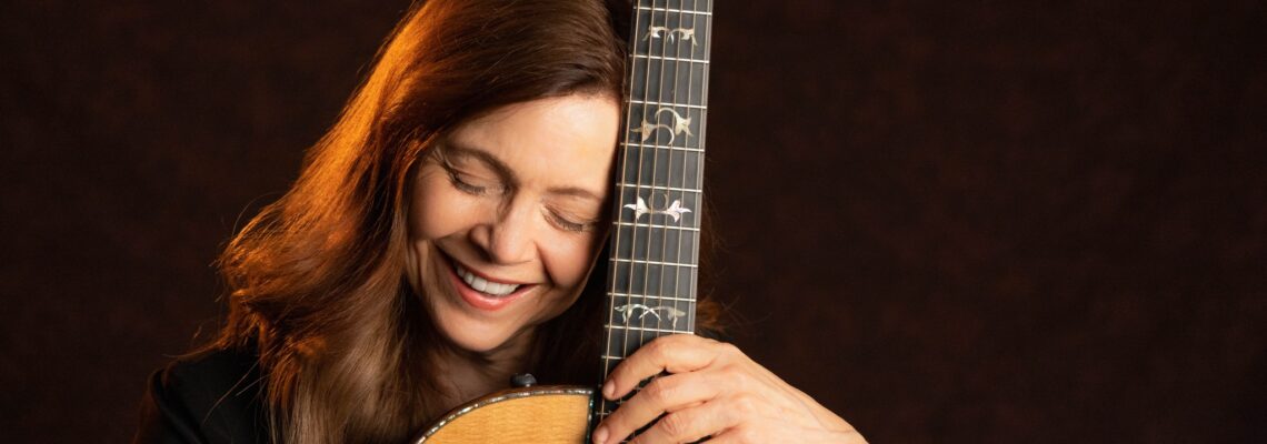 a middle-aged white woman with long brown hair, smiling, eyes closed, posing with a guitar